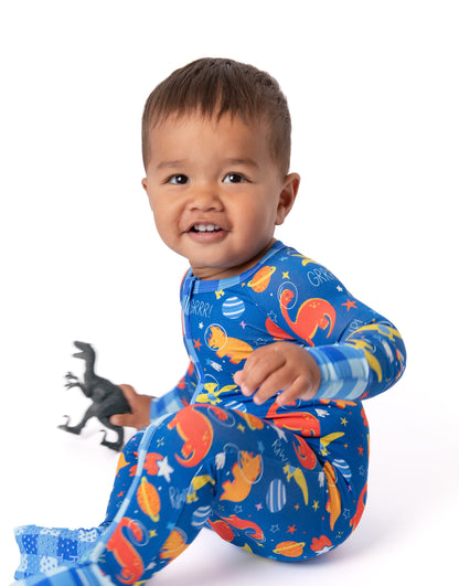 A smiling toddler sitting on a white studio background wearing a blue long-sleeve footed pajama set with a colorful dinosaur and space print, holding a small black toy dinosaur.