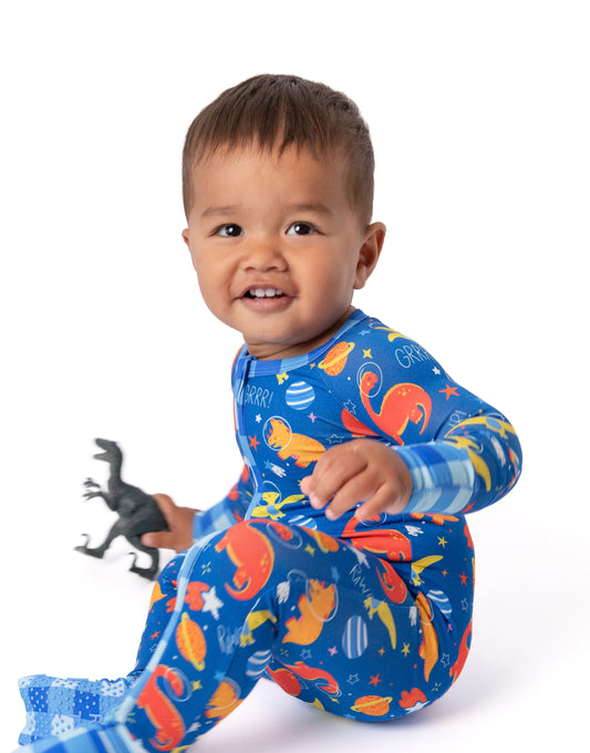 A smiling toddler sitting on a white studio background wearing a blue long-sleeve footed pajama set with a colorful dinosaur and space print, holding a small black toy dinosaur.