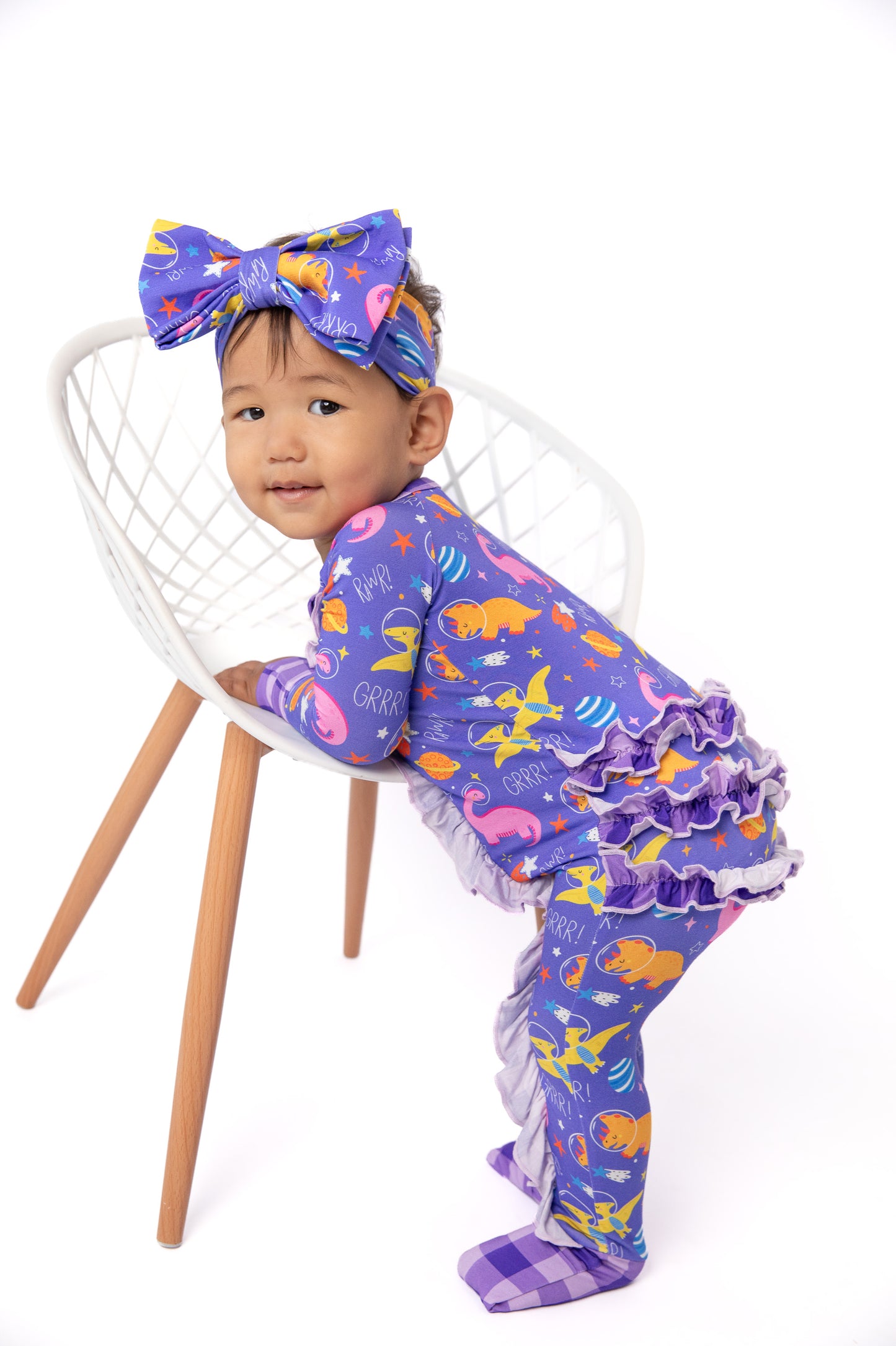 A toddler wearing a purple footed pajama set with a colorful ocean-themed print, a matching bow headband, and ruffled trim, posing on a white chair against a white background.
