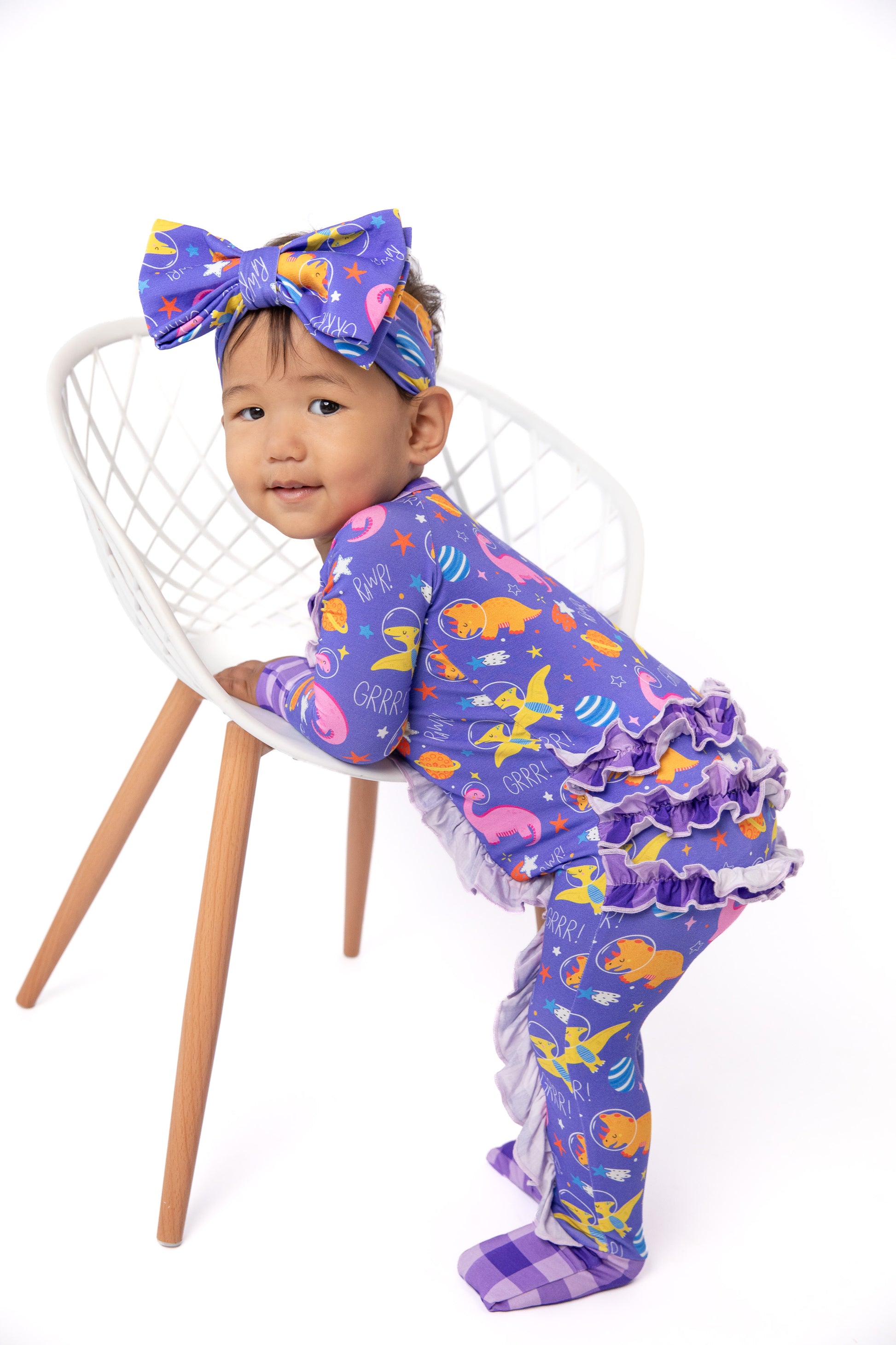 A toddler wearing a purple footed pajama set with a colorful ocean-themed print, a matching bow headband, and ruffled trim, posing on a white chair against a white background.