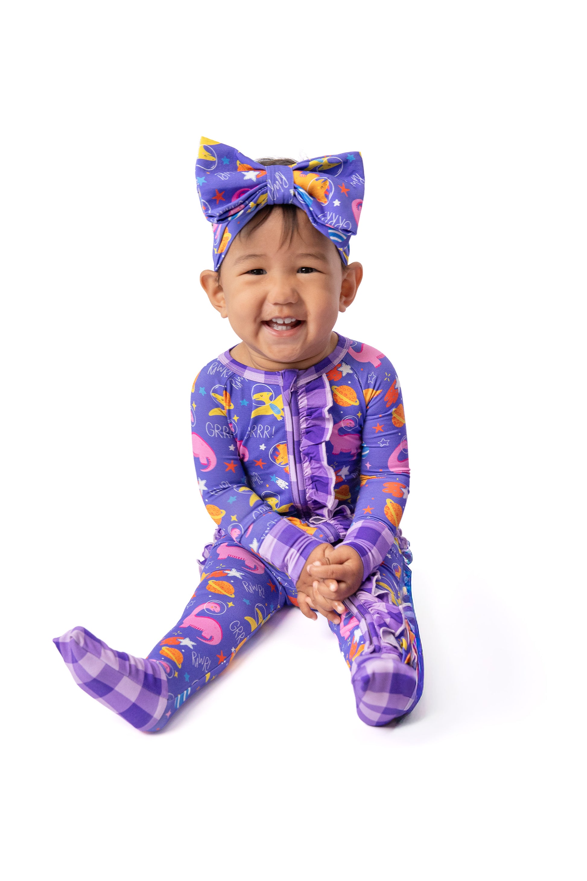 A smiling toddler sitting on a white background wearing a purple full-body onesie with a colorful flamingo print, matching bow headband, and purple plaid socks.