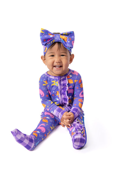 A smiling toddler sitting on a white background wearing a purple full-body onesie with a colorful flamingo print, matching bow headband, and purple plaid socks.