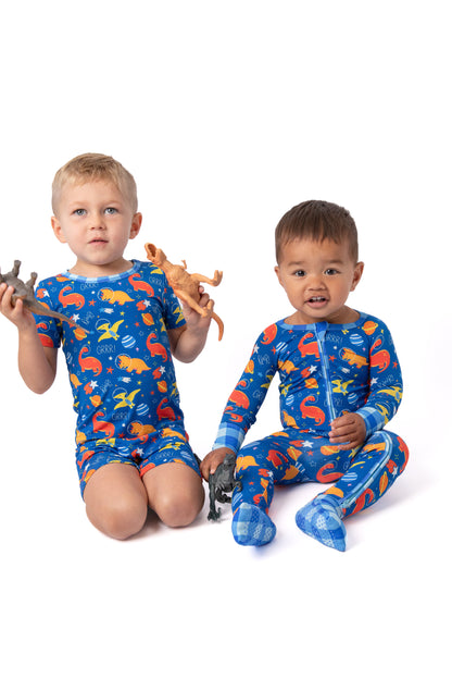 Two young children wearing matching blue dinosaur-print pajamas sit on a white studio background, each holding small toy dinosaurs.