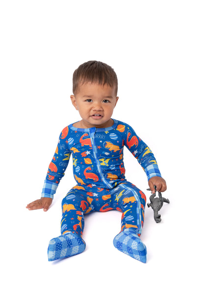A baby sitting on a white background wearing a blue footed pajama top and pants set with a colorful dinosaur print and a blue-and-white checkered collar and cuffs. The baby is holding a small gray toy.
