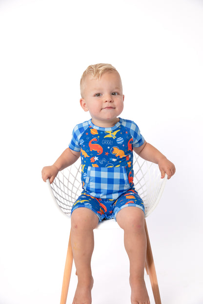 A toddler sitting on a white wire chair wearing a blue and white gingham short-sleeve romper with colorful dinosaur and fish prints, photographed against a clean white studio background.