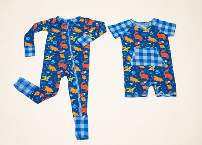 Two children's long-sleeve pajamas laid flat on a light beige background. One is a full-body one-piece with a blue gingham cuff and hem, and the other is a short romper with a blue gingham collar and sleeve, both featuring a blue background print with orange, yellow, and white dinosaurs and stars.