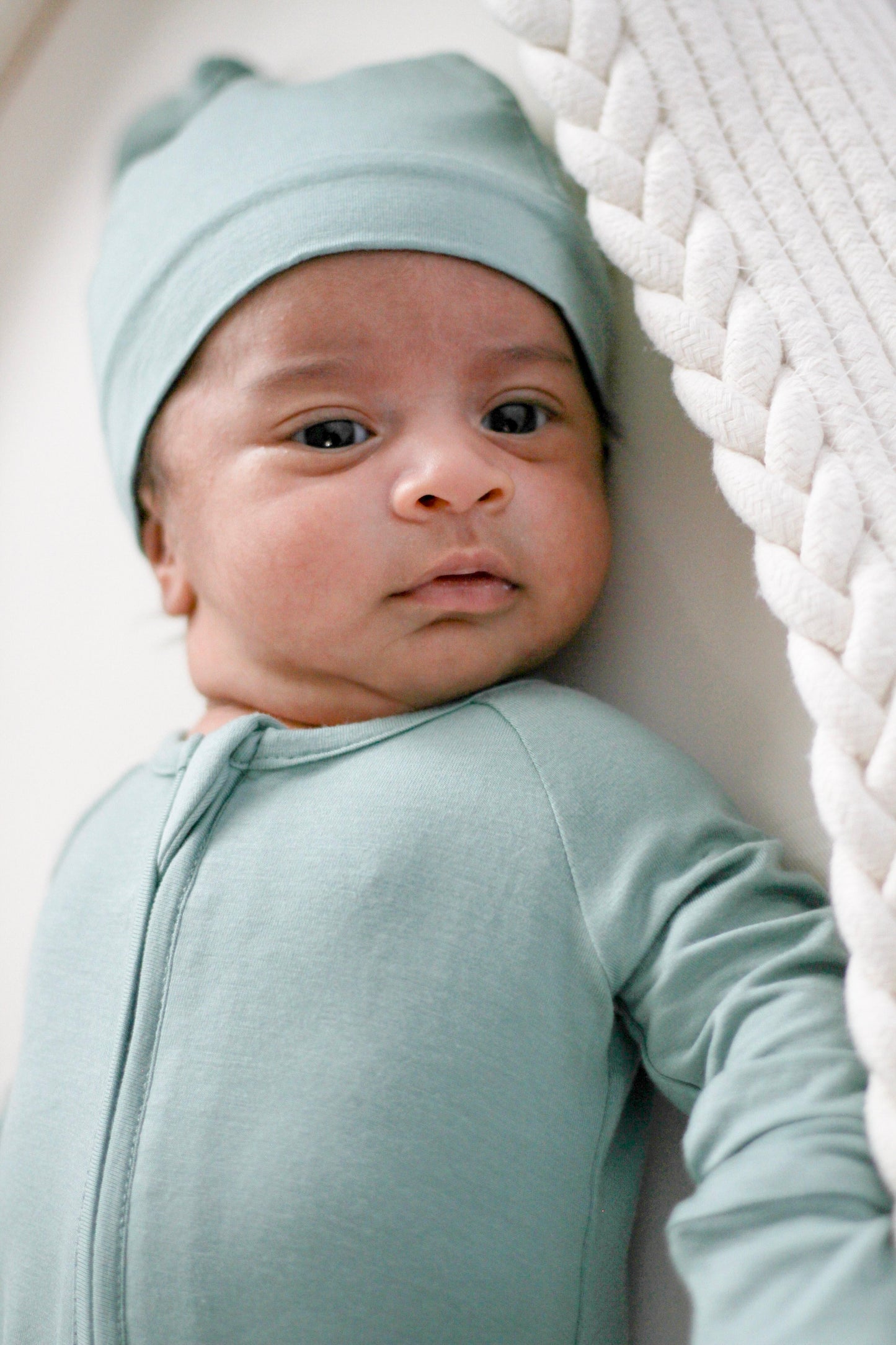 Baby wearing a light blue outfit and hat, sitting against a white textured background