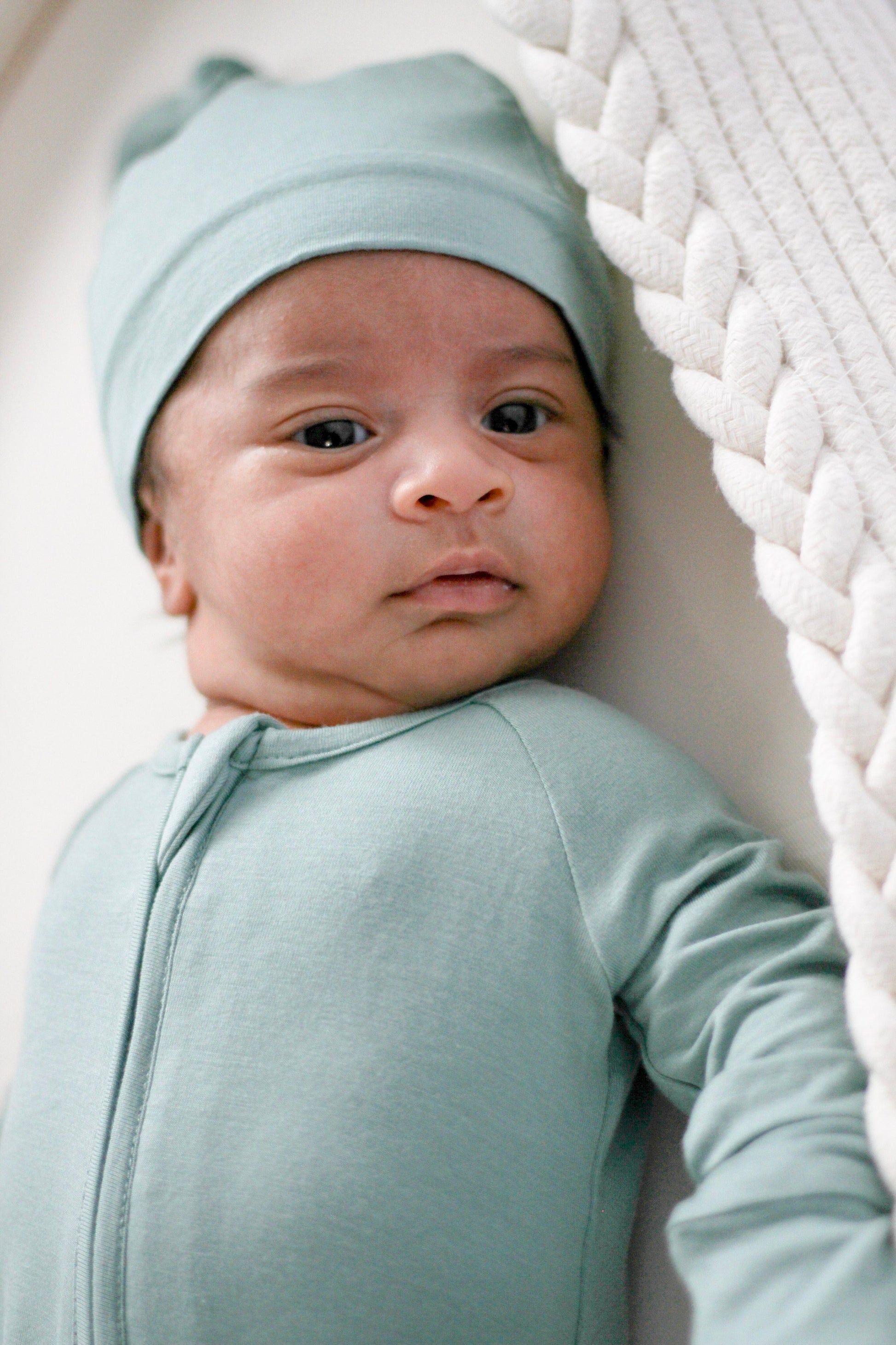 Baby wearing a light blue outfit and hat, sitting against a white textured background