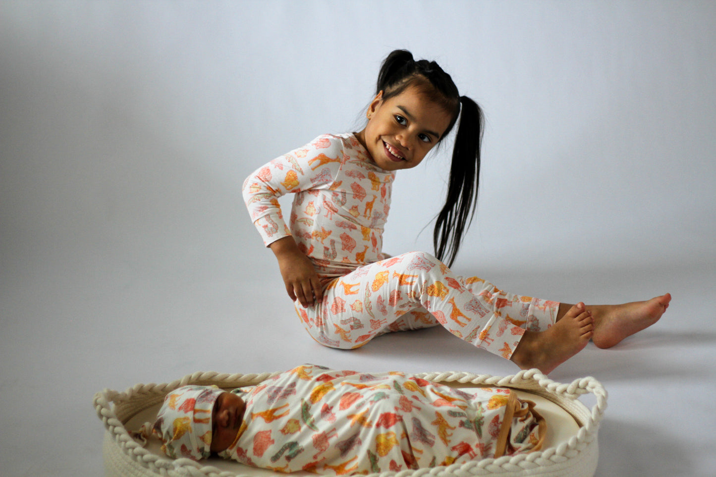 Child wearing a patterned outfit sitting on a white surface with a baby lying in a basket below.