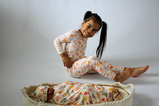 Child wearing a patterned outfit sitting on a white surface with a baby lying in a basket below.
