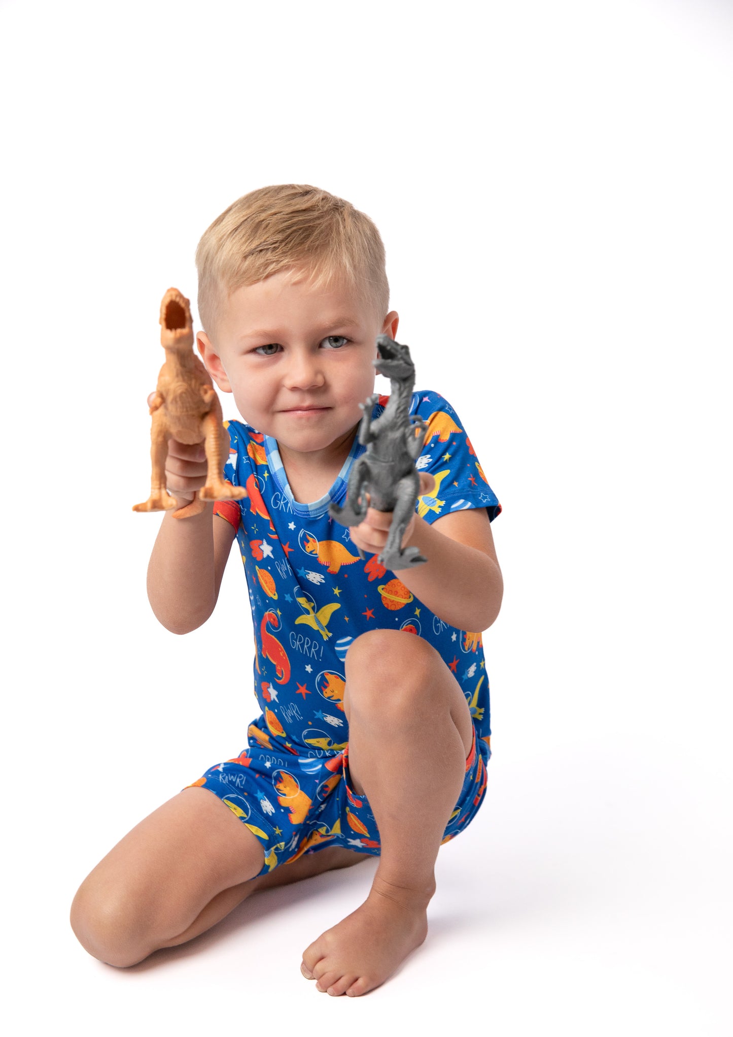 A young child kneeling on a white studio background, wearing a blue short-sleeve one-piece pajama with a colorful dinosaur print and holding two small dinosaur toys.
