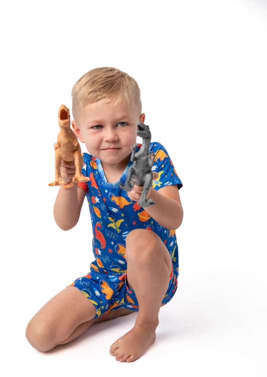 A young child kneeling on a white studio background, wearing a blue short-sleeve one-piece pajama with a colorful dinosaur print and holding two small dinosaur toys.