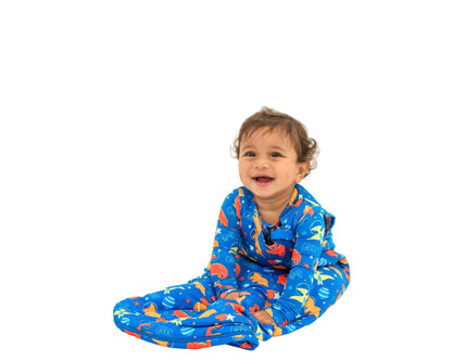A smiling toddler sitting cross-legged on a clean white studio background, wearing a bright blue footed pajama or swaddle with an allover orange and yellow dinosaur print.