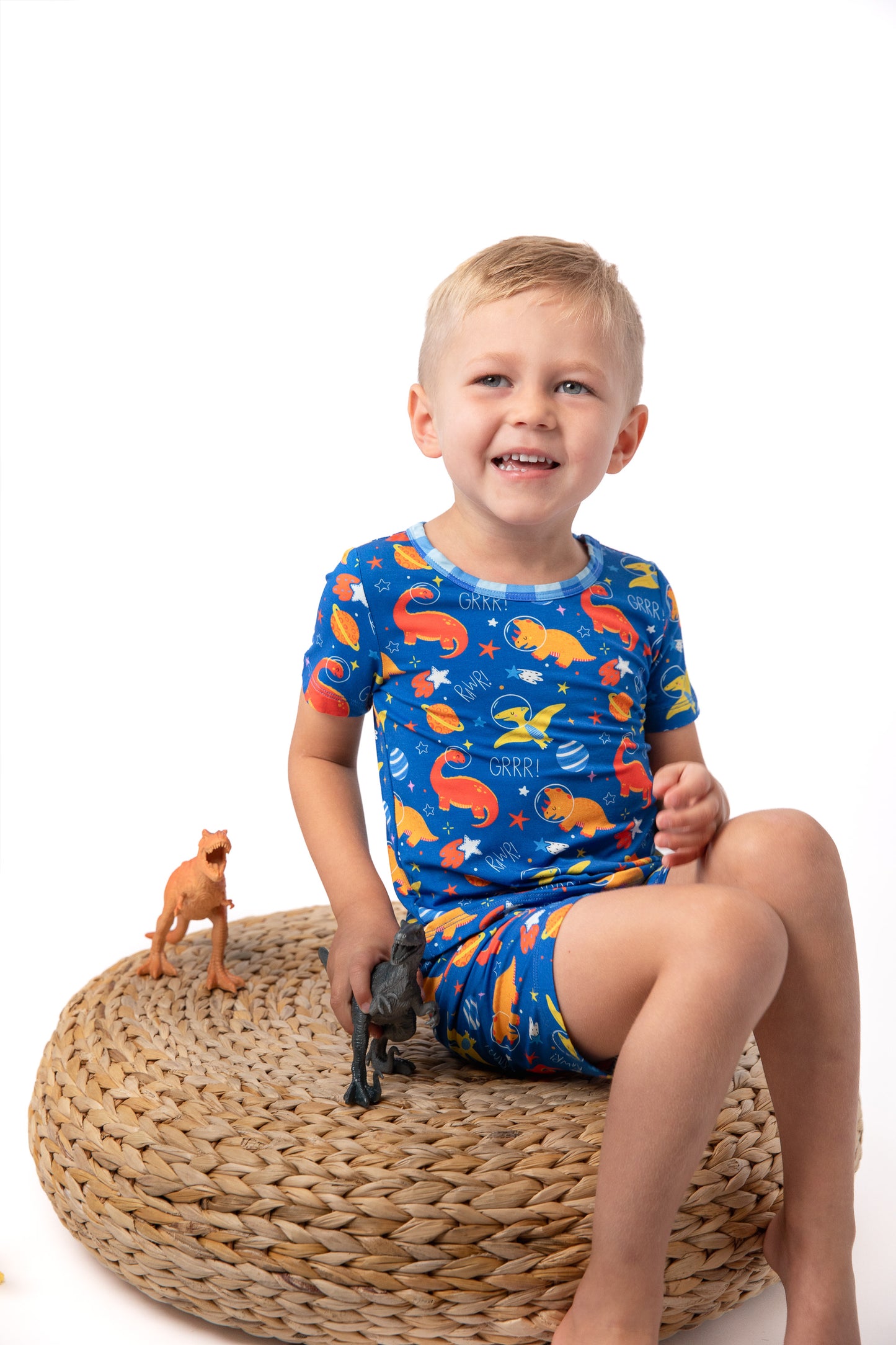 a boy in space dinosaur blue short pajamas sitting on a cushion with dinosaurs 