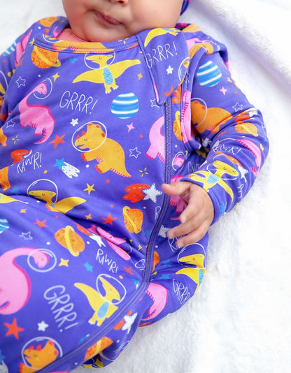A baby wearing a purple zip-up footed sleeper with a colorful dinosaur and space-themed print, lying on a white textured blanket.