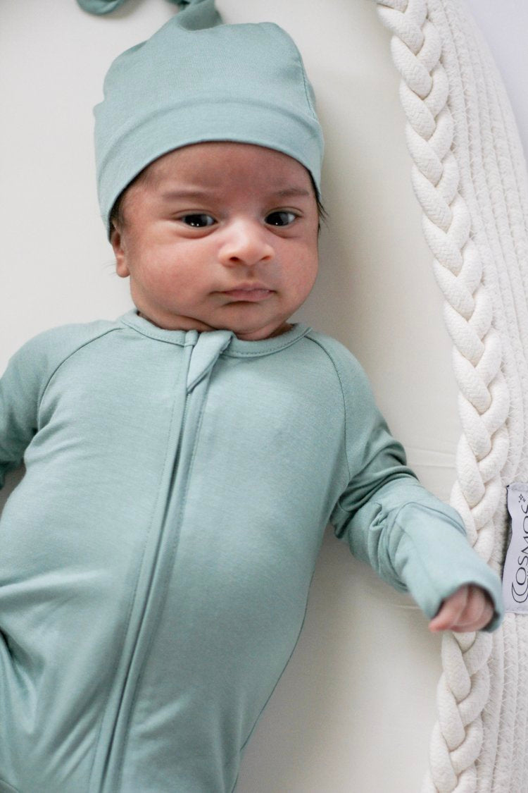 Baby in a light blue outfit and hat lying on a white surface with a textured white blanket.