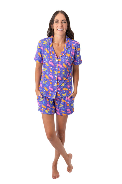 A woman modeling a matching short-sleeve button-up pajama shirt and shorts set with a colorful tropical print on a solid white background.