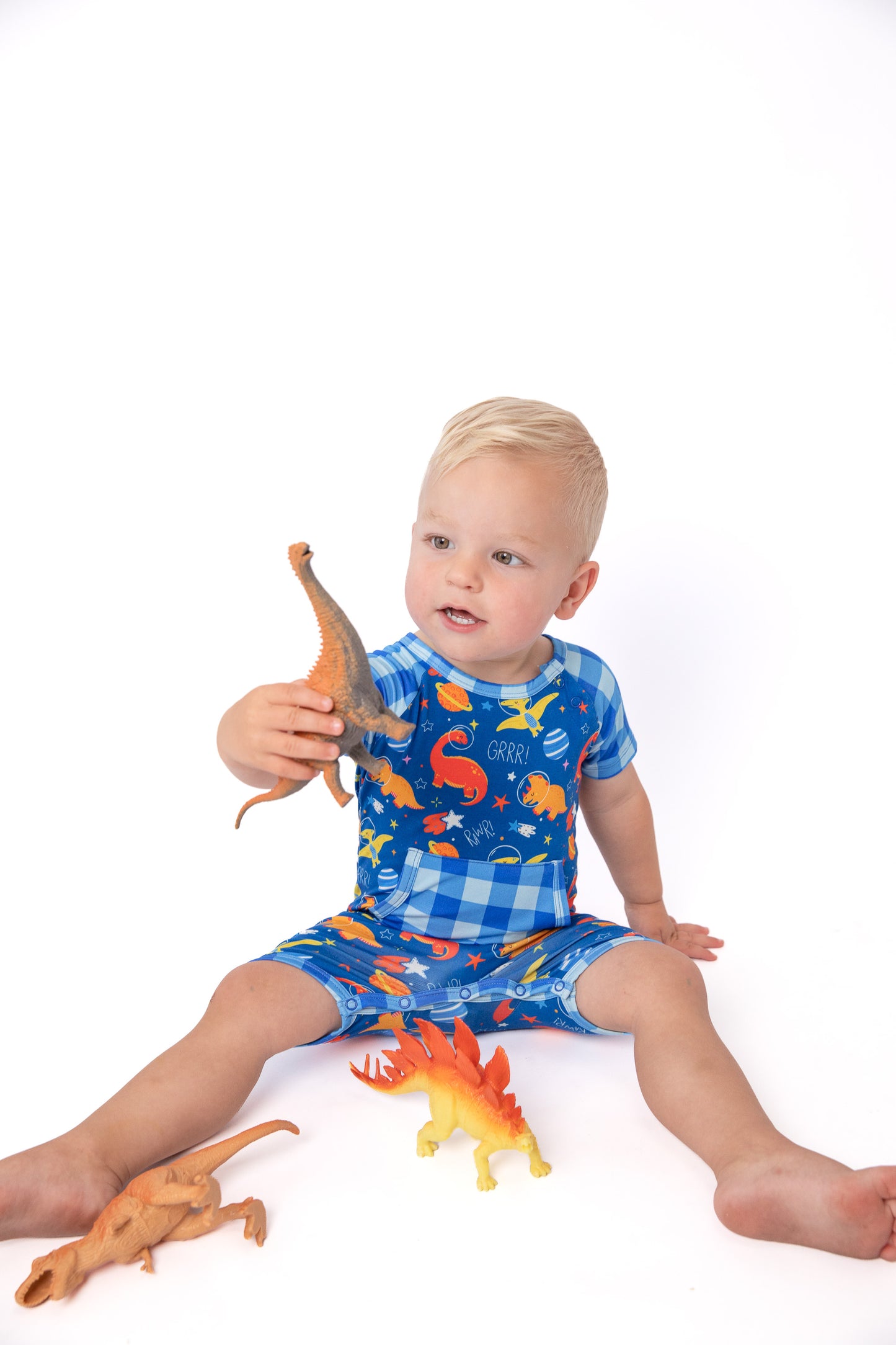 A toddler sitting on a white studio background wearing a blue dinosaur-print pajama romper, holding a small dinosaur toy.