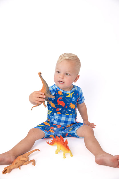 A toddler sitting on a white studio background wearing a blue dinosaur-print pajama romper, holding a small dinosaur toy.