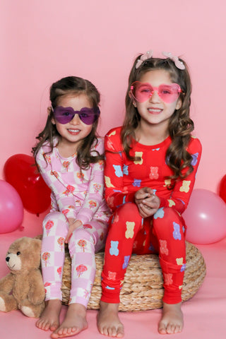 Two children in colorful pajamas with heart-shaped sunglasses on a pink background.
