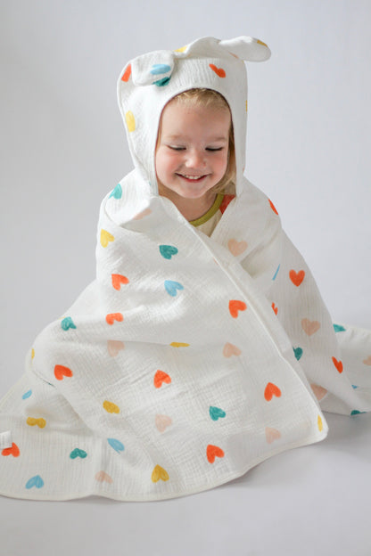 Child wrapped in a colorful heart-patterned hooded towel on a plain background