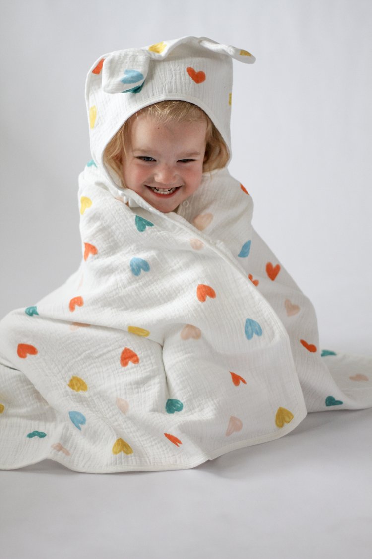 Child wrapped in a colorful heart-patterned towel with a hood on a plain background