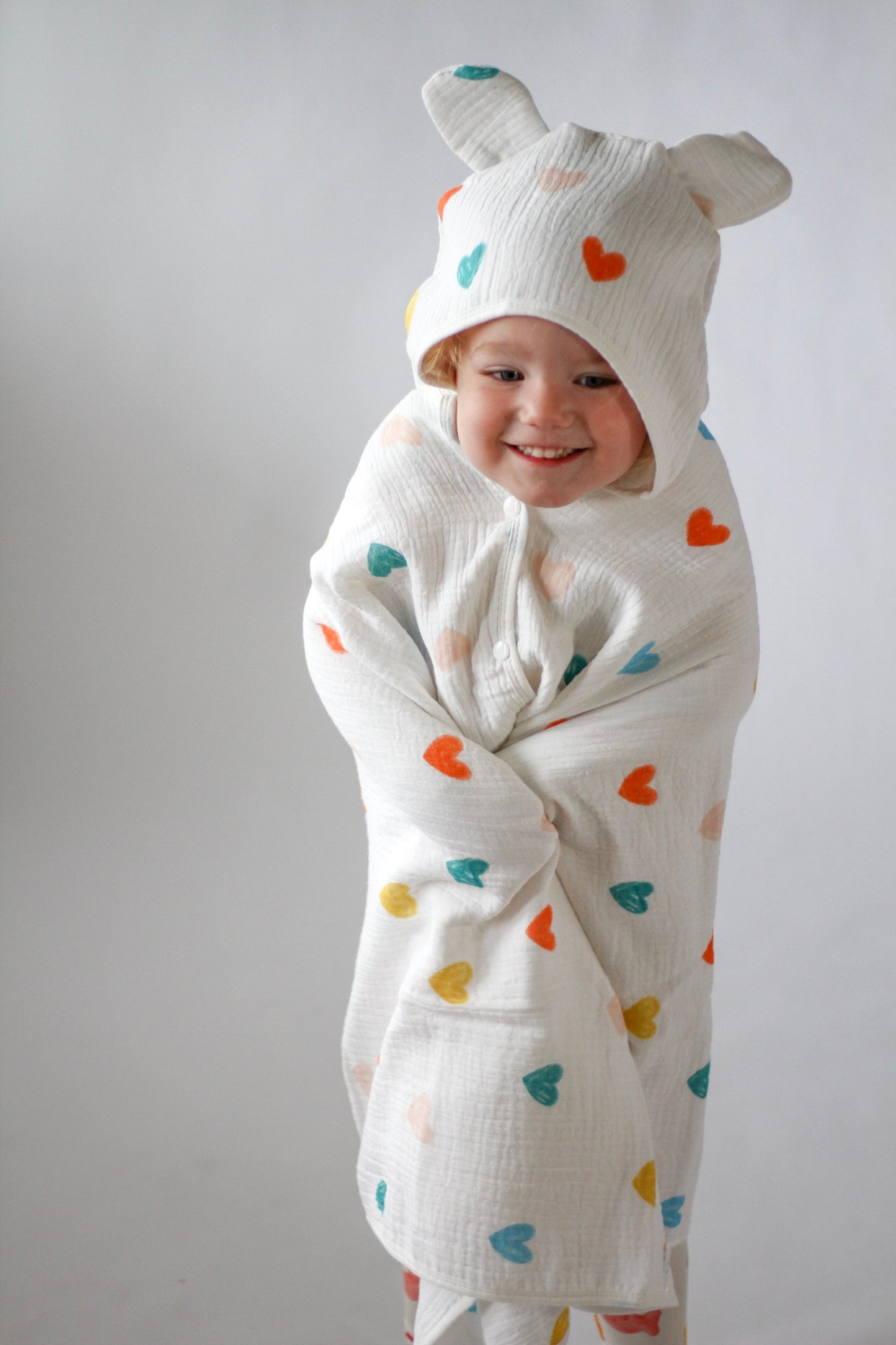 Child wearing a white onesie with colorful heart patterns and a matching hat on a plain background