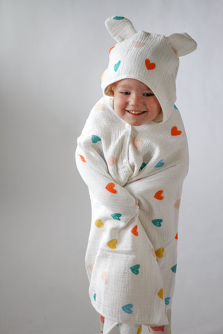 Child wearing a white onesie with colorful heart patterns and a matching hat on a plain background