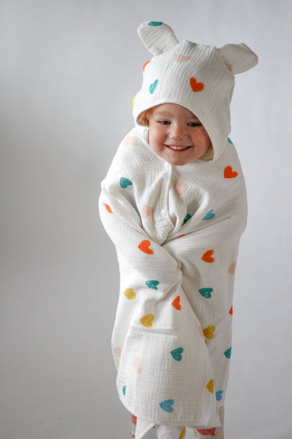 Child wearing a white onesie with colorful heart patterns and a matching hat on a plain background