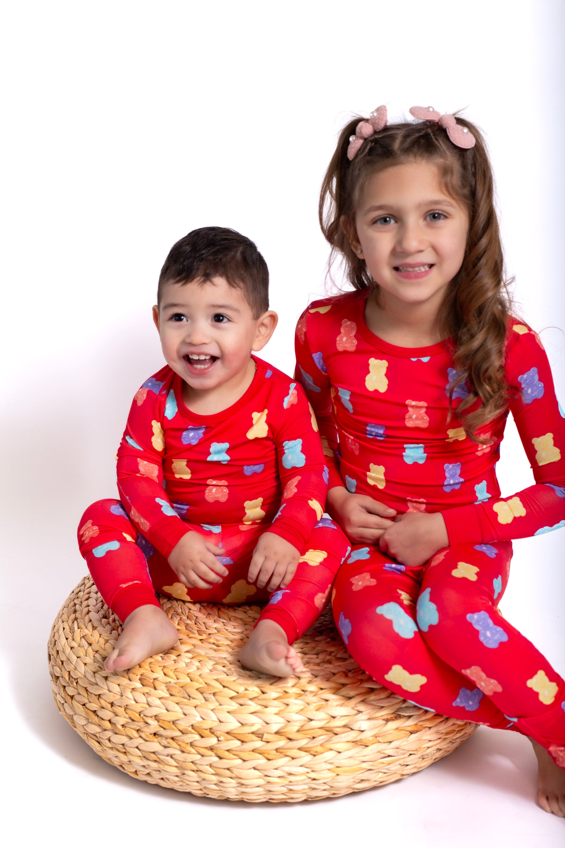 Two children in red pajamas with bear patterns sitting on a woven stool against a white background.