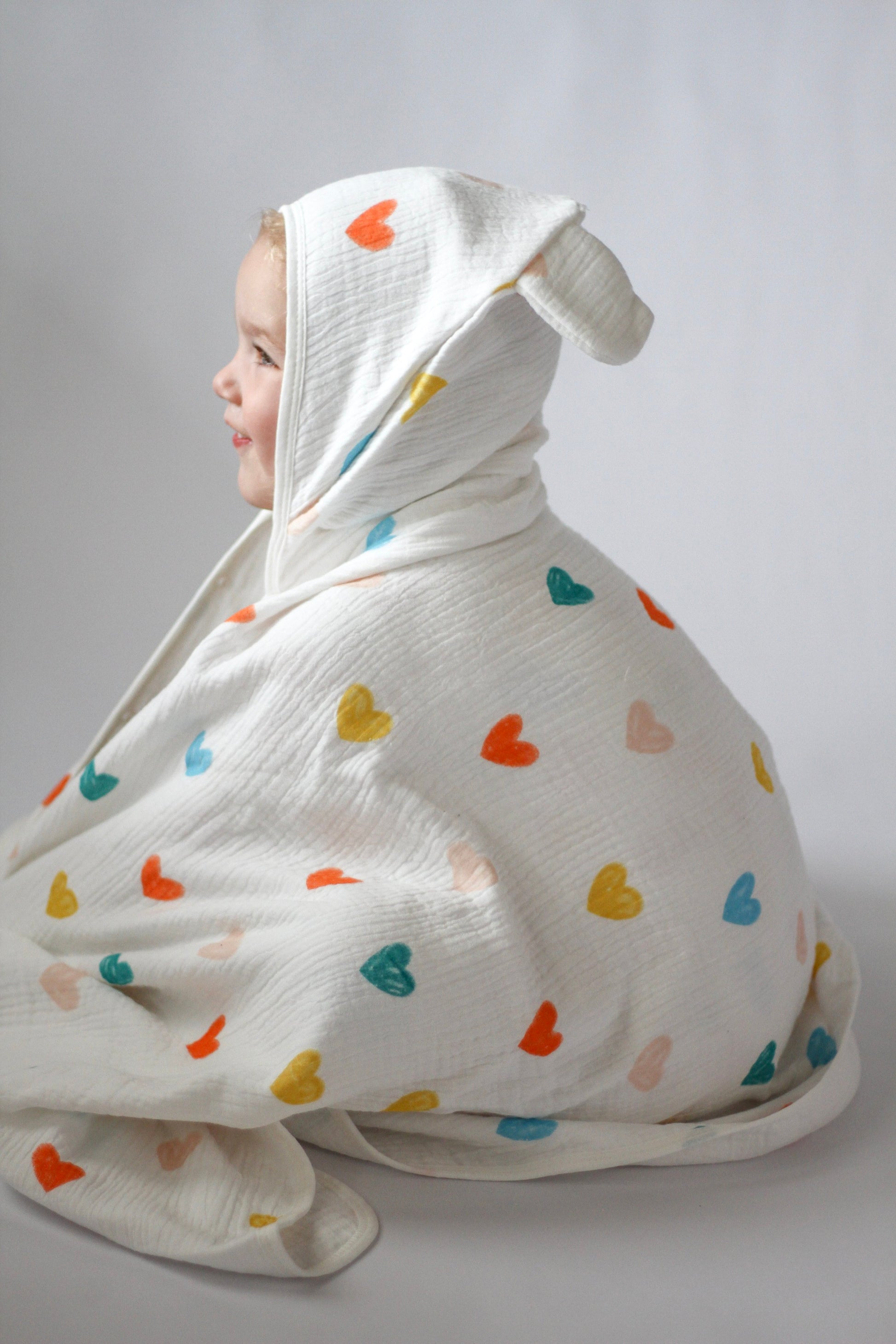 Child wrapped in a white blanket with colorful heart patterns on a plain background