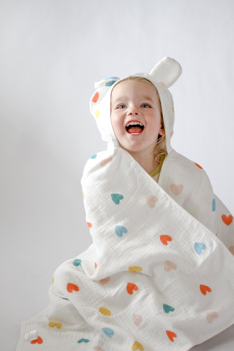 Child wrapped in a white hooded blanket with colorful heart patterns on a plain background