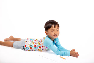 Child lying on a white surface with a pencil, wearing a colorful shirt and patterned pants.