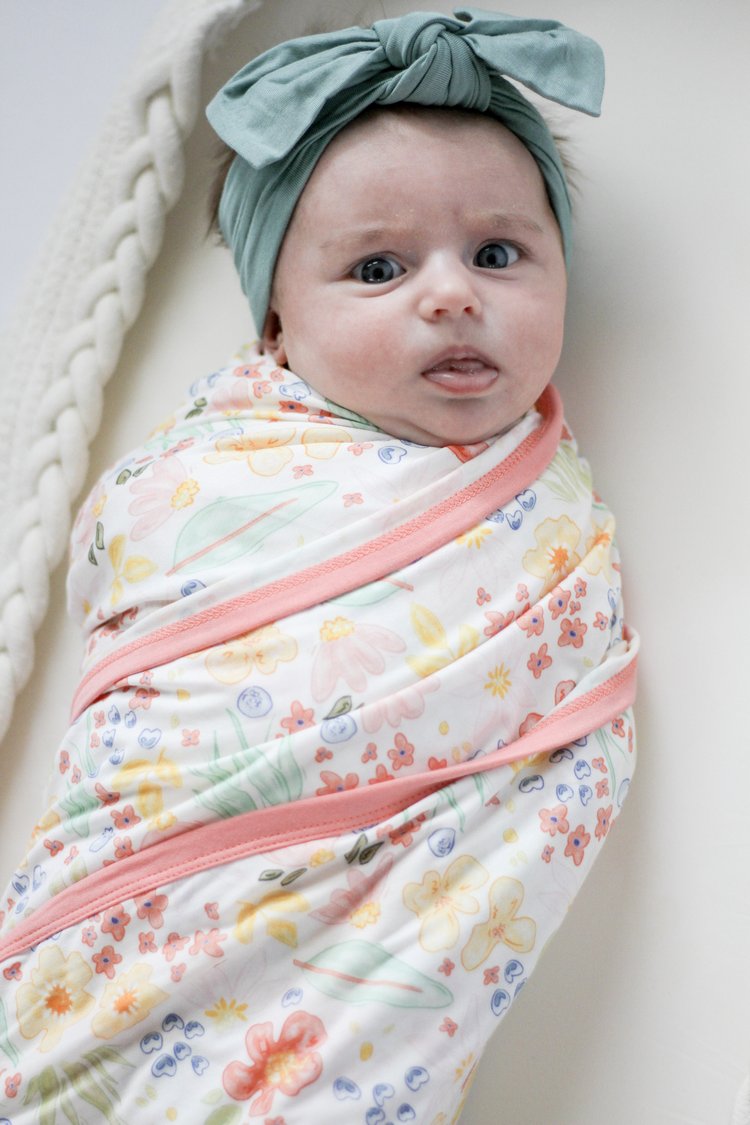Baby wrapped in a floral swaddle with a green headband on a neutral background