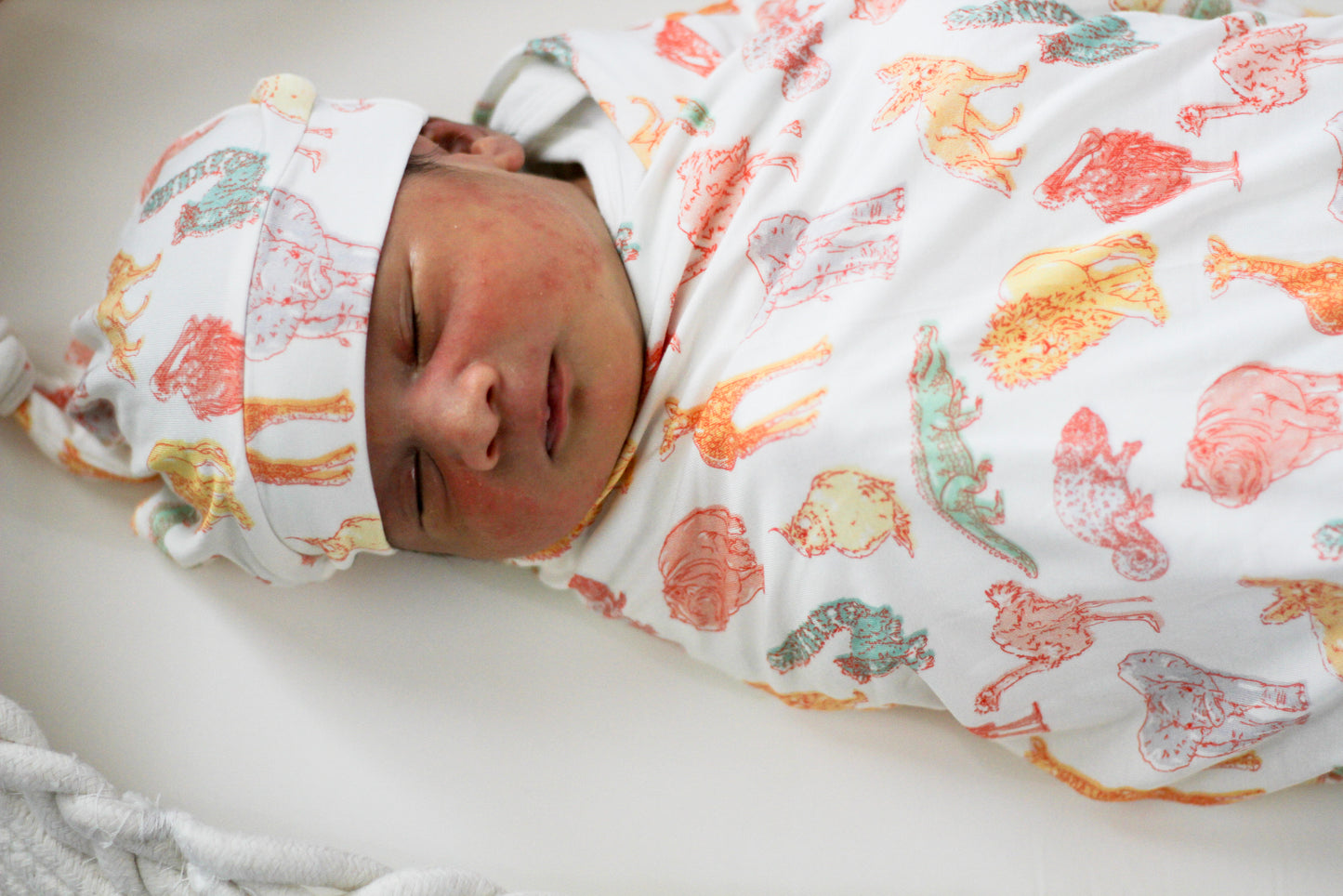 Newborn baby swaddled in a colorful blanket with animal patterns on a white surface.