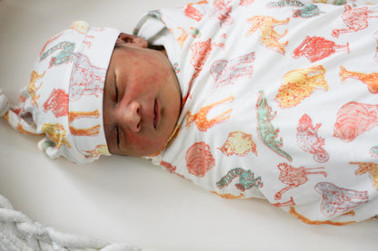 Newborn baby swaddled in a colorful blanket with animal patterns on a white surface.