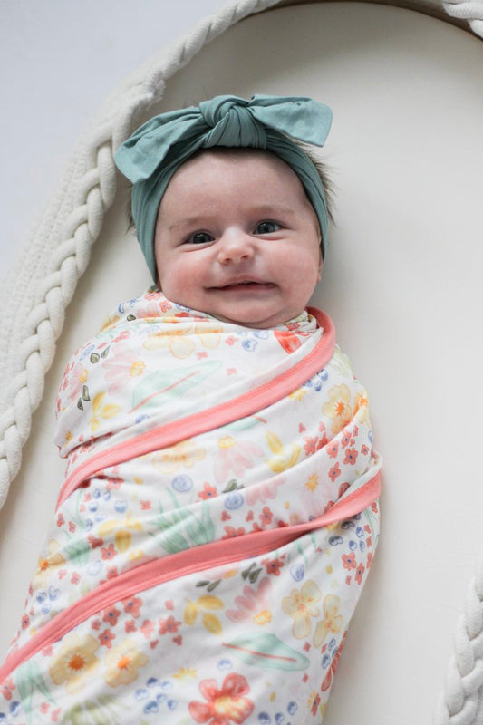 Baby swaddled in a floral blanket with a headband, lying on a white surface.