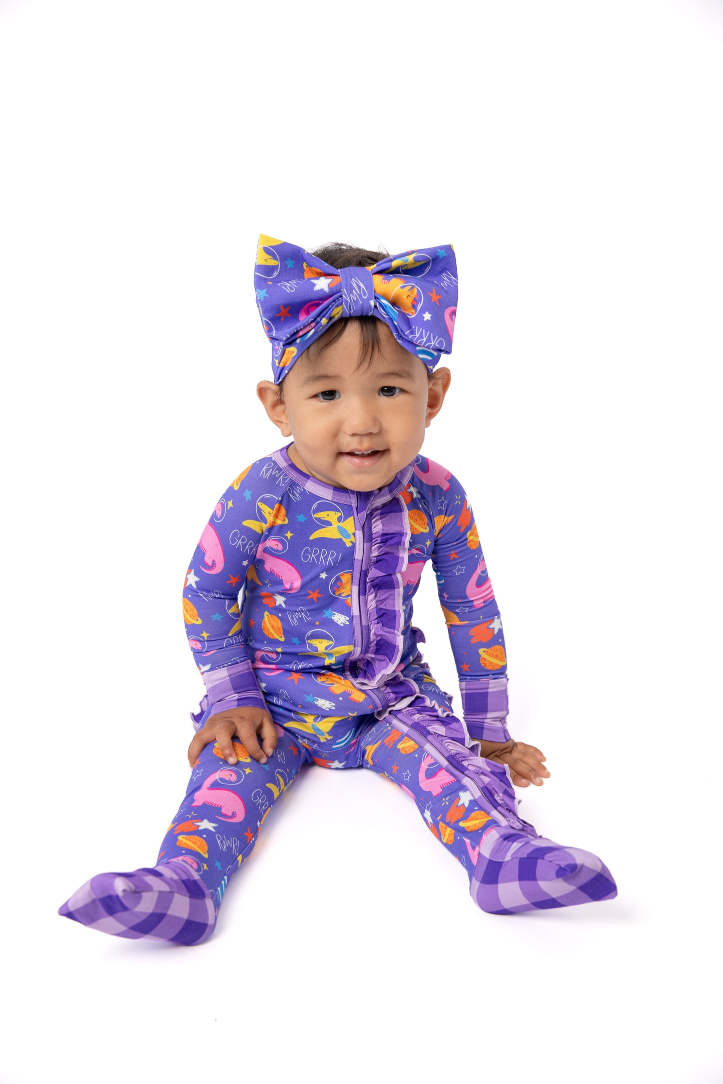 A toddler sitting on a white studio background wearing a purple footed pajama set with a matching bow headband. The pajamas feature a colorful print of flamingos, fish, and carrots on a purple plaid base.