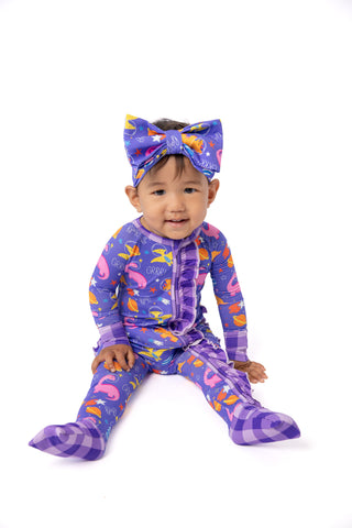 A toddler sitting on a white studio background wearing a purple footed pajama set with a matching bow headband. The pajamas feature a colorful print of flamingos, fish, and carrots on a purple plaid base.