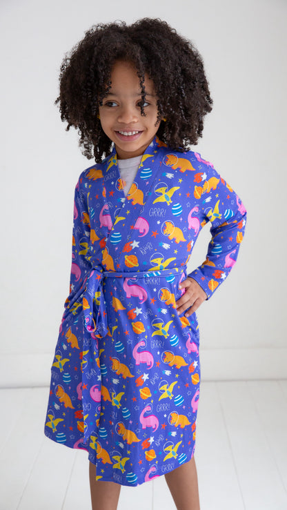 A young girl with curly dark hair smiling while wearing a long-sleeve wrap robe with a colorful mermaid and fish pattern on a purple background.