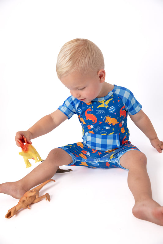 A toddler sitting on a white studio floor wearing a blue dinosaur-print short-sleeve romper and matching shorts, holding a small yellow dinosaur toy.