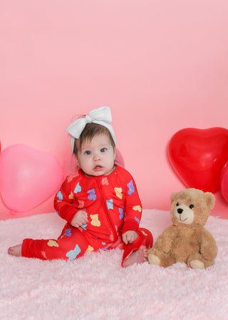 Baby in a red outfit with colorful patterns sitting on a pink surface with balloons and a teddy bear.
