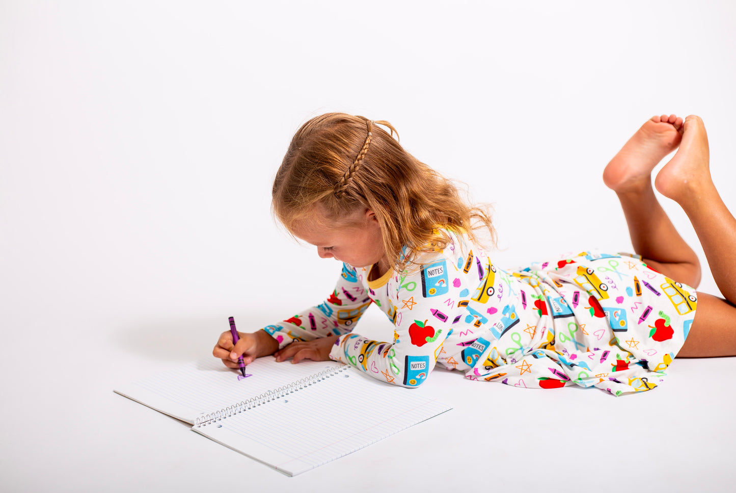 Child in pajamas with a colorful pattern, lying on a white floor and drawing in a notebook.