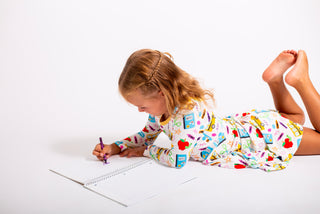 Child in pajamas with a colorful pattern, lying on a white floor and drawing in a notebook.