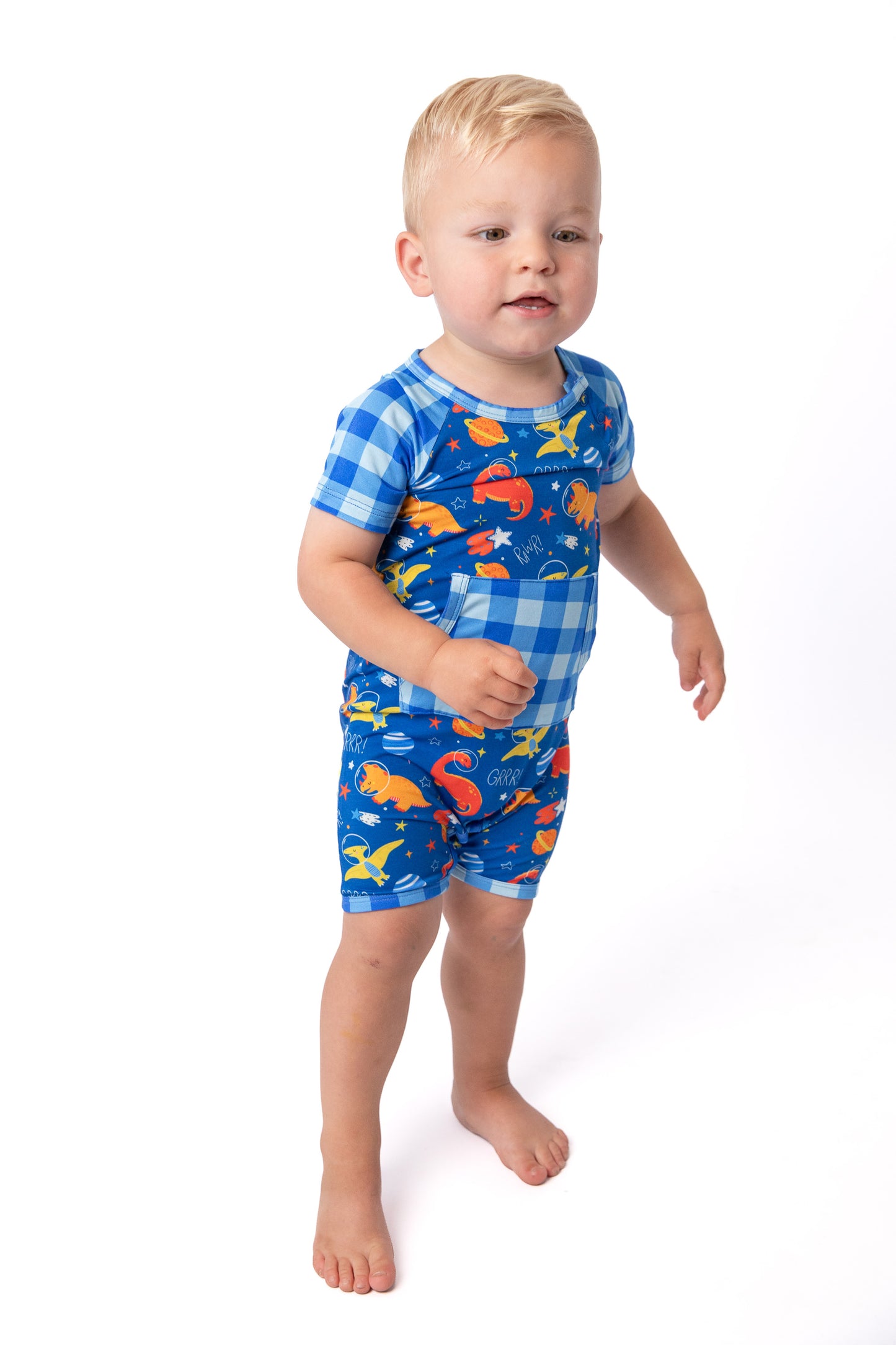 A toddler standing against a white studio background wearing a short-sleeve one-piece swimsuit with a blue and white gingham pattern on the upper body and legs, and an ocean-themed print with orange, yellow, and blue fish on a blue background on the lower half.