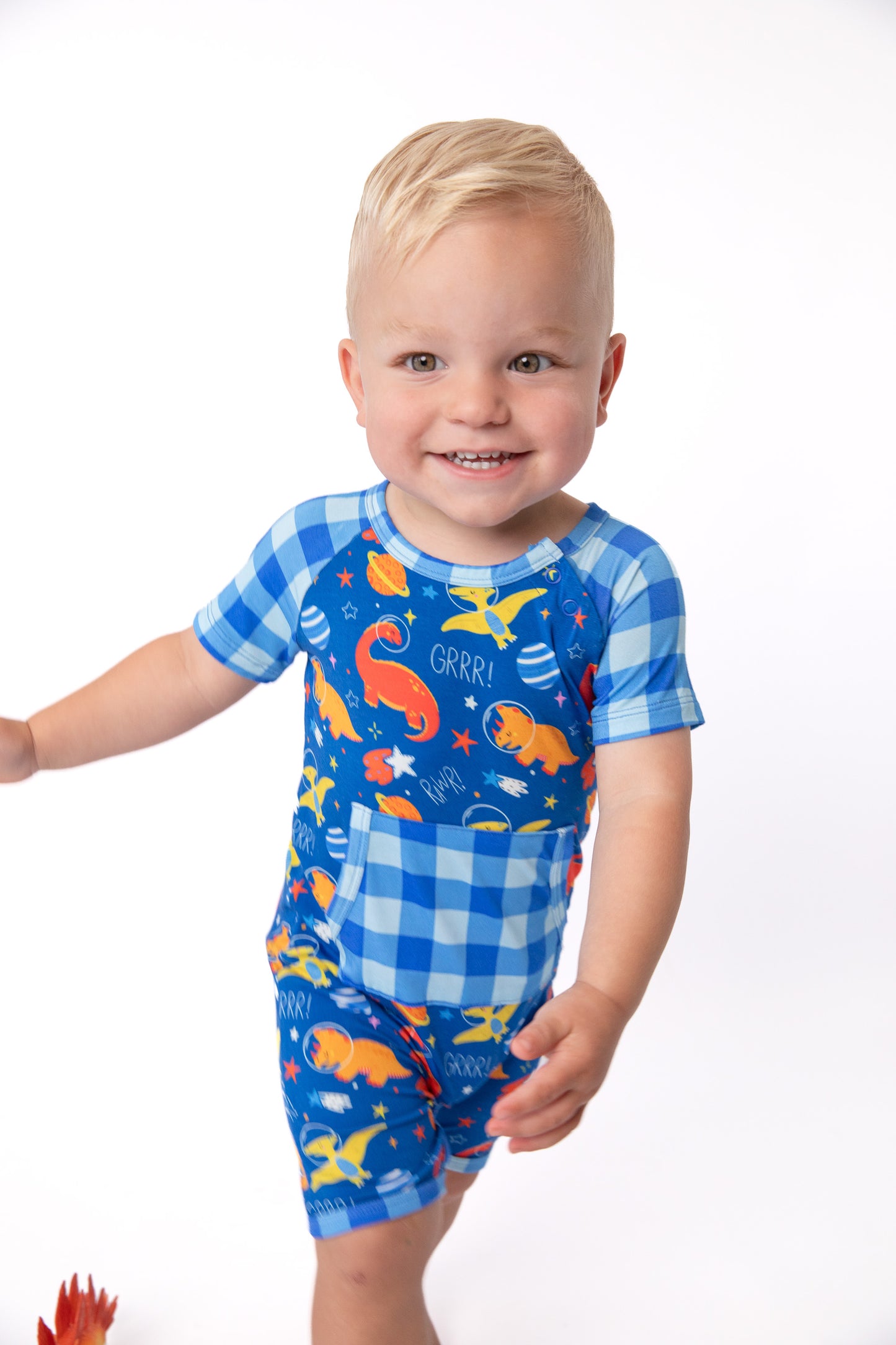 A smiling toddler wearing a blue short-sleeve romper with a gingham pattern and colorful dinosaur print, standing against a plain white background.