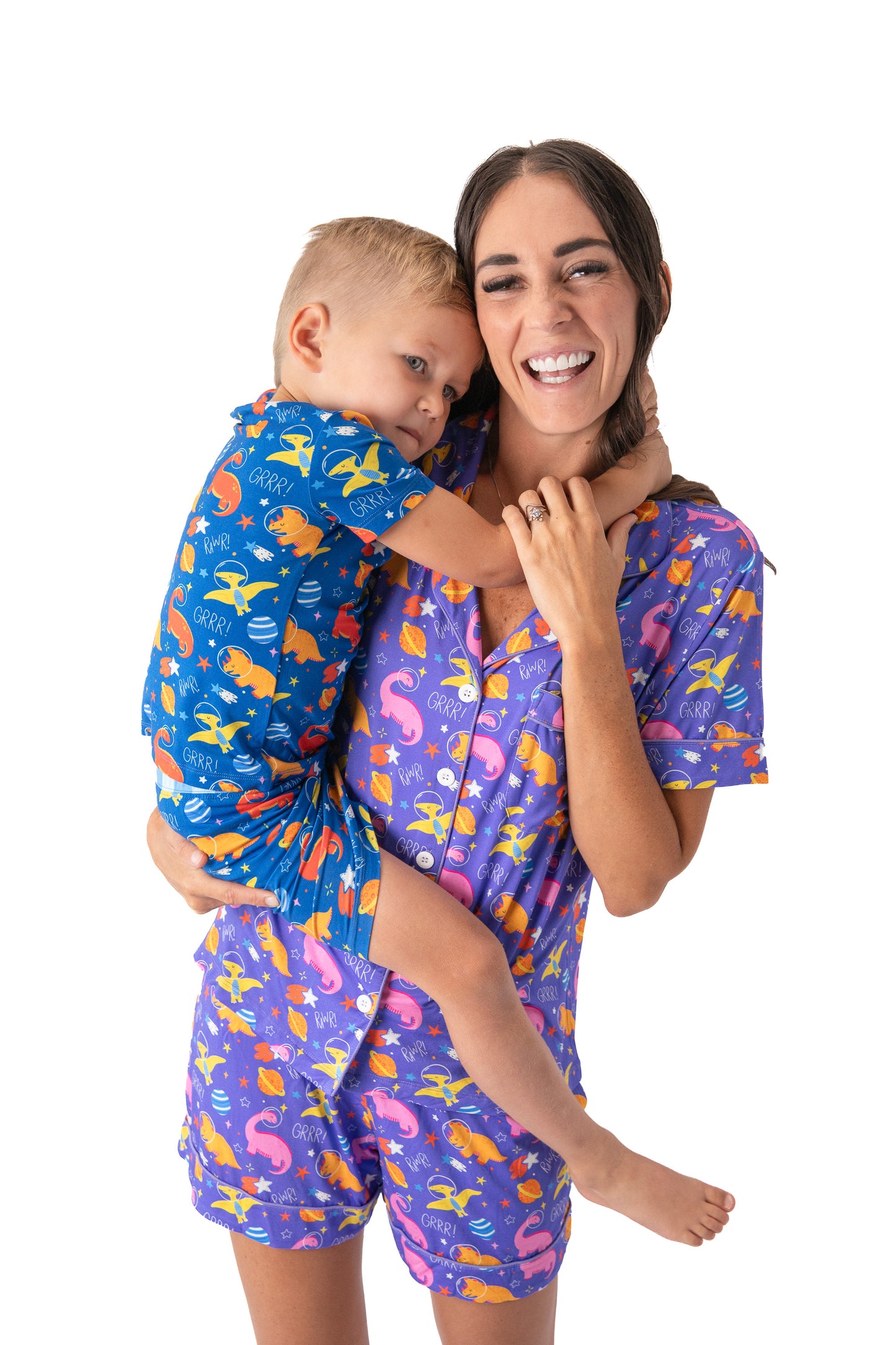 A woman and a young boy wearing matching pajamas with a colorful dinosaur print on a blue and purple background, posed against a solid white studio background.