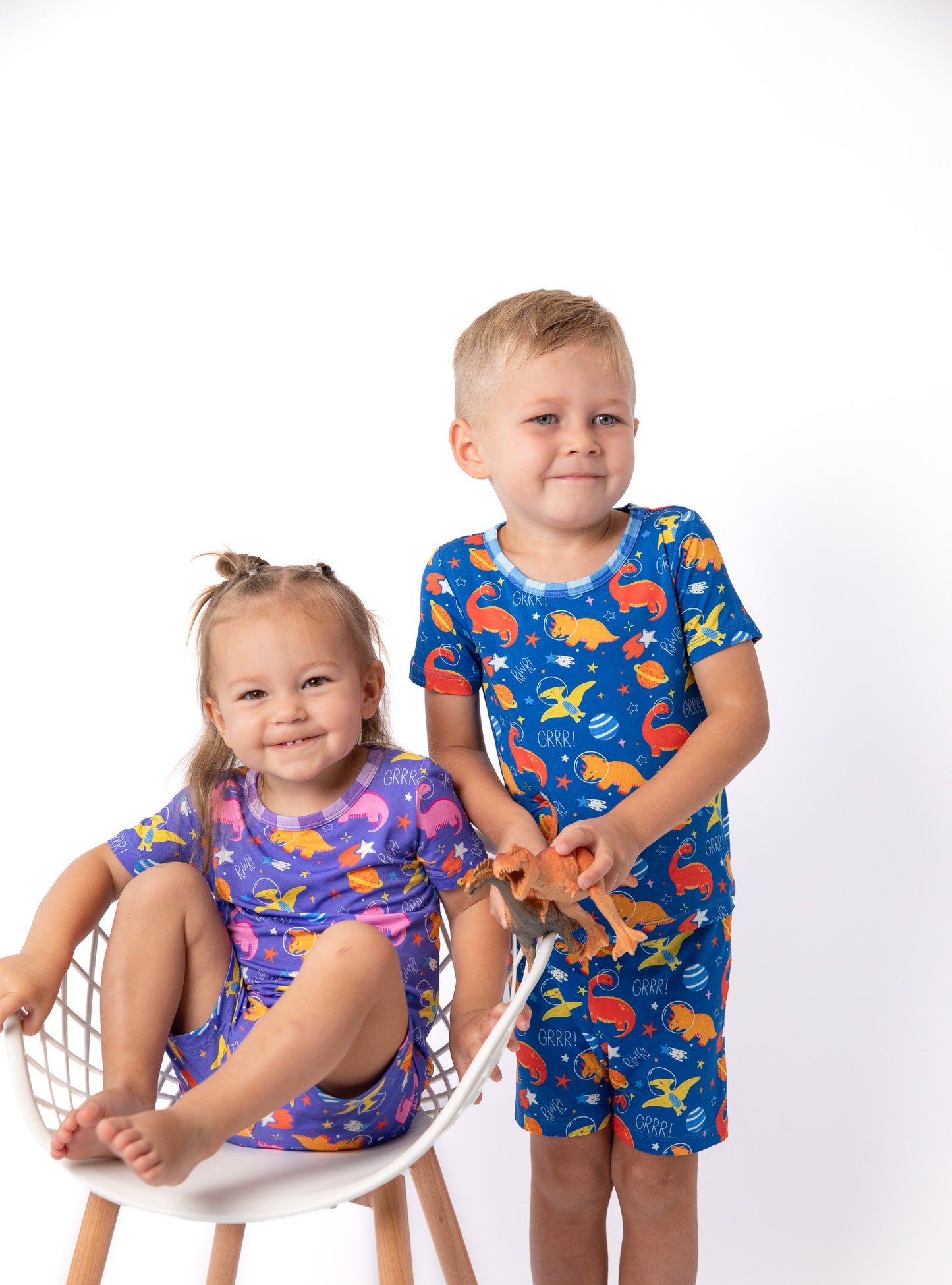 Two children modeling matching pajamas against a white studio background. The girl sits on a white chair wearing a purple pajama set with pink, orange, and yellow fish print.