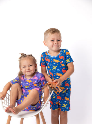 Two children modeling matching pajamas against a white studio background. The girl sits on a white chair wearing a purple pajama set with pink, orange, and yellow fish print.