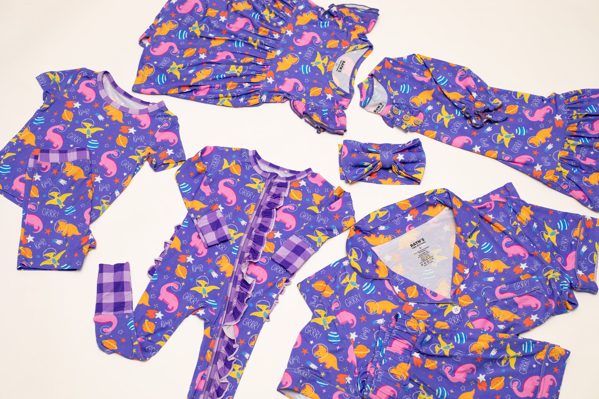 A coordinated baby clothing set laid flat on a light cream background, including a purple long-sleeve romper, matching long-sleeve top and pants, a matching headband, and a zip-front footless sleeper. The fabric has an allover mermaid print in pink, orange, yellow, and blue, with some plaid trim details.